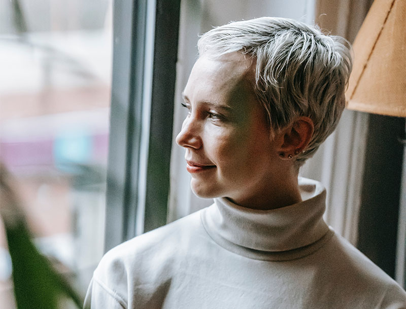 Frau mit kurzen, weißen Haaren und weißem Rollkragenpullover blickt lächelnd aus dem Fenster.
