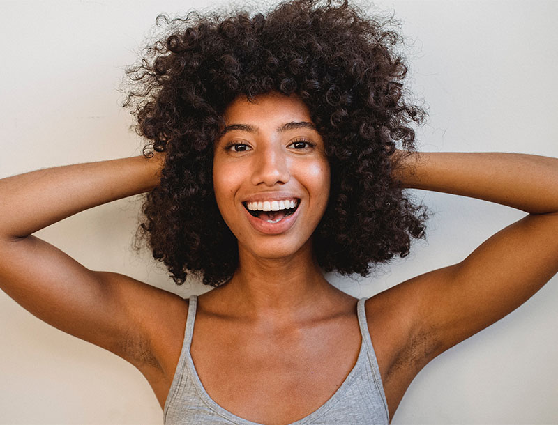 Lachende Frau mit lockigen Haaren hält Hände hinter dem Kopf.